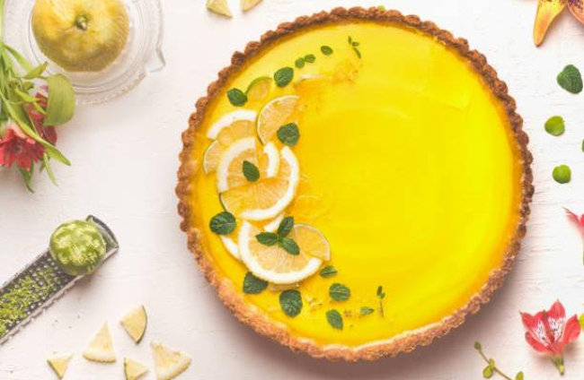 Yellow lemon tart topped with fresh lemon and lime slices on white table background with citrus ingredients and flowers, top view. Traditional french cuisin