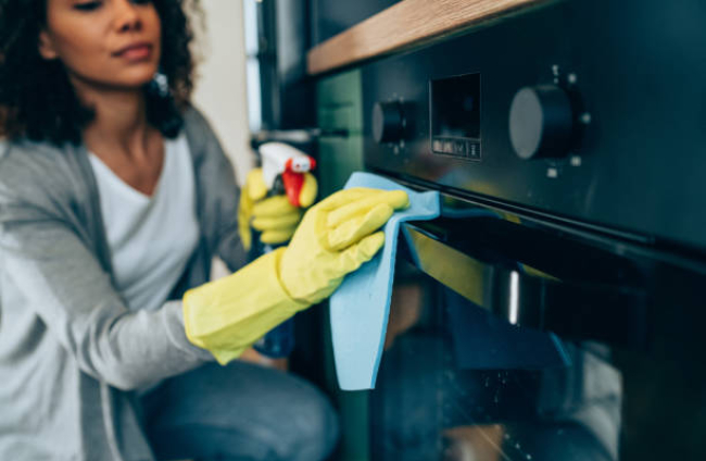 Shot of young woman cleaning the outside of an oven. Woman cleaning with spray disinfectant and gloves at home