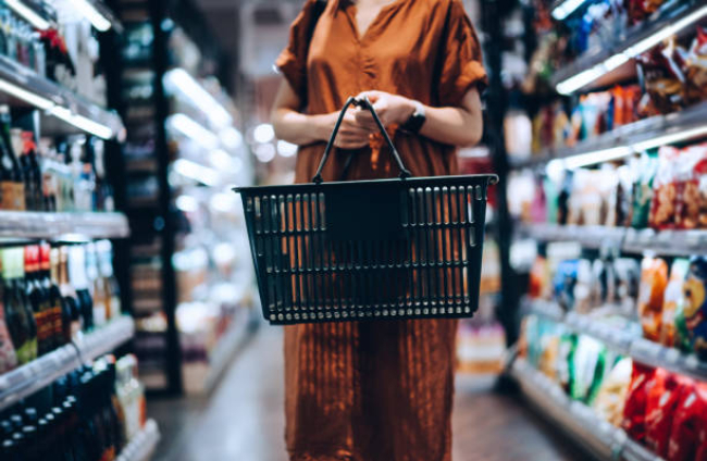 Cropped shot of young woman carrying a shopping basket, standing along the product aisle, grocery shopping for daily necessities in supermarke