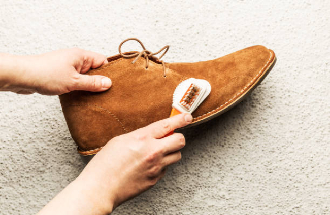 Hands cleaning men's camel suede desert shoe (boot) with a brush. Footwear maintenance captured from above (top view). Grey concrete background