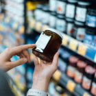 Close up of young woman grocery shopping in a supermarket. Standing by the aisle, holding a bottle of marmalade, reading the nutritional label and checking ingredients at the bac